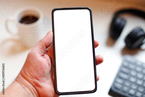 Hand Holding Smartphone With Blank White Screen In Modern Workspace With Coffee And Headphones with copy-space