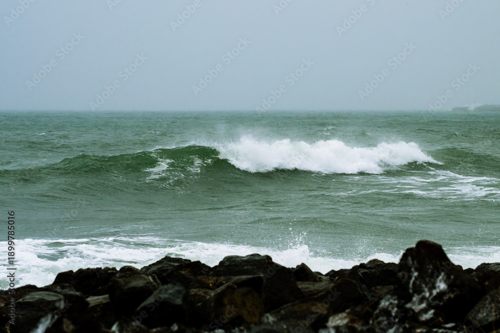 Fototapeta premium turbulent green ocean waves crashing onto a rocky shore, sending white foam and spray into the air.