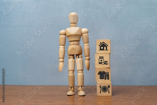 Photography A wooden doll stands next to stacked blocks with icons for housing, healthcare, clothing, and food