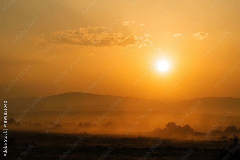 Obraz premium Hazy sunset panorama: warm orange tones, smoky clouds crossing the sun, serene distant hills