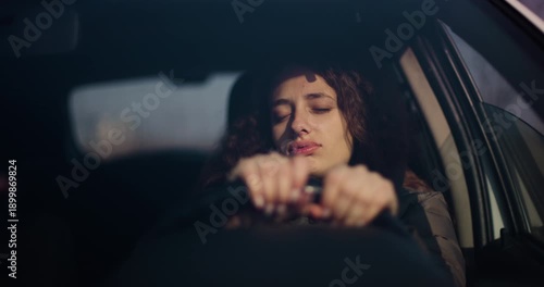 A young woman with visible braces looks sleepy as she drives her car during the evening. The reflection of the sunset can be seen in the window.