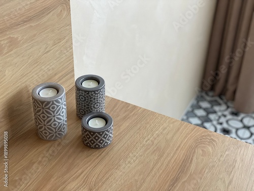 Minimalist still life of three decorative ceramic candle holders with geometric patterns placed on a wooden surface.Neutral gray tones,natural light, and simple interior background.