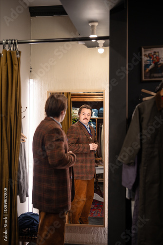 Wallpaper Mural Elegant man with mustaches buying blazer in vintage men's clothing boutique, looking in reflection checks outfit and appearance. Male customer in second hand store fits on wool jacket in fitting room. Torontodigital.ca
