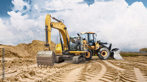 Heavy machinery is at work on sandy ground, leaving deep tracks. A large pile of sand is visible in the background indicating ongoing construction activities