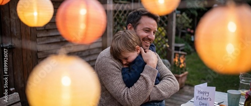 Heartwarming Portrait of a Happy Father Hugging His Little Son during Outdoor Celebration. Cozy Evening Backyard Party with Glowing Paper Lanterns and Greeting Card for Father's Day. Panoramic Banner 