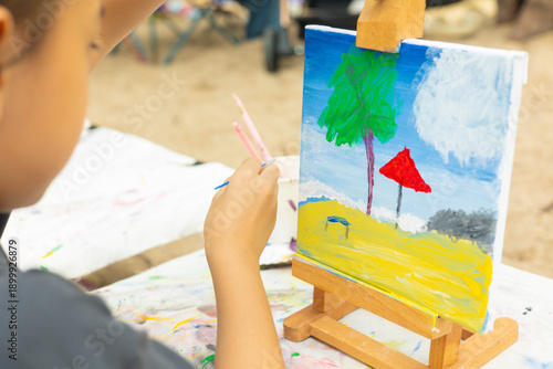 Close-up and selective focus of young girl painting a palm tree and sea with beautiful color on canvas. This shows creativity of a child's artistic hobby in a natural, outdoor beach setting.