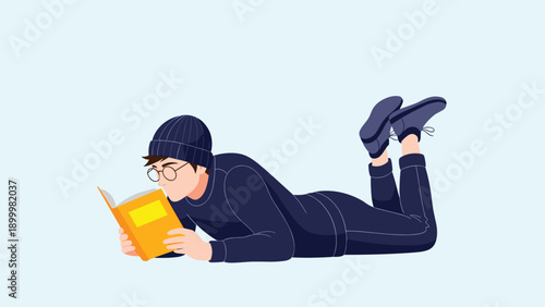 Young man wearing a beanie and glasses, comfortably lying on his stomach and engrossed in reading a book, portraying quiet leisure and learning.