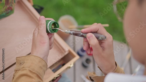 Hands painting green ornament outdoors, asian artist applies precise brush strokes on miniature surface, wooden easel and open jars on grass, palette with acrylic colors, focused meditative mood,