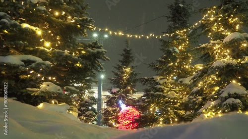 Christmas decoration of city square. Winter evening with snowfall. Christmas trees are decorated with shining bulbs and garlands. Large red lamp on snow in form of Christmas ball. New Year Landscape