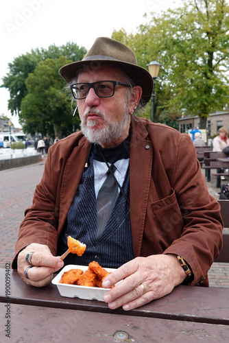 Elderly man with a beard, wearing a brown hat, glasses and a brown jacket. He's seated outdoors at a picnic table, holding a takeout container of Dutch fried fish with a fork. 