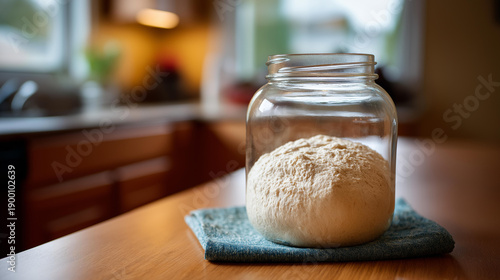 Homemade yeast starter for sourdough bread in glass jar, kitchen table with living culture for baking preparation, artisan bread making with natural leavening agent display,