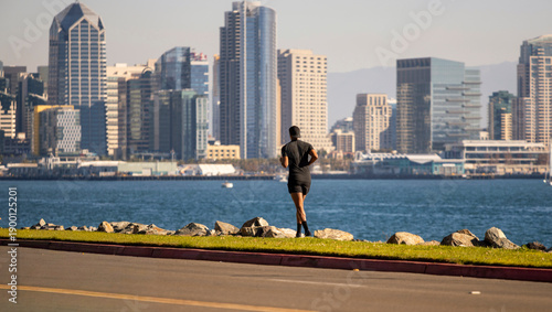 A physically fit man dressed in black jogging on a trail with the City of San Diego in the background