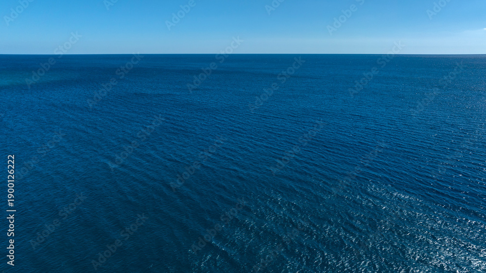 Fototapeta premium A vast, high-angle view of a deep blue sea with gentle ripples on the surface, meeting a clear, light blue horizon under a bright sky.