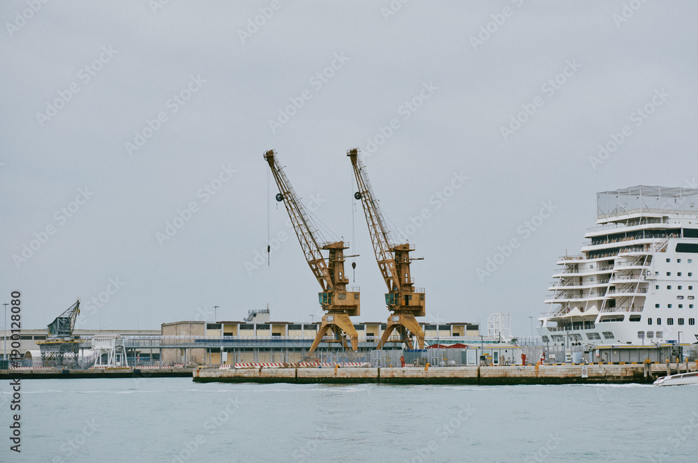 Obraz premium Two yellow harbor cranes standing idle on a concrete quay beside a large white cruise ship under grey sky