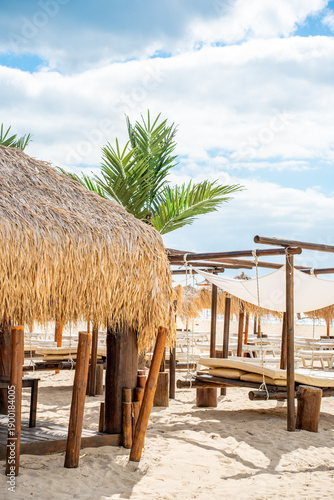 Beach huts made of straw stand along the shoreline. Sun loungers are arranged under the huts. The sky is blue with some clouds, and the sand is light and soft