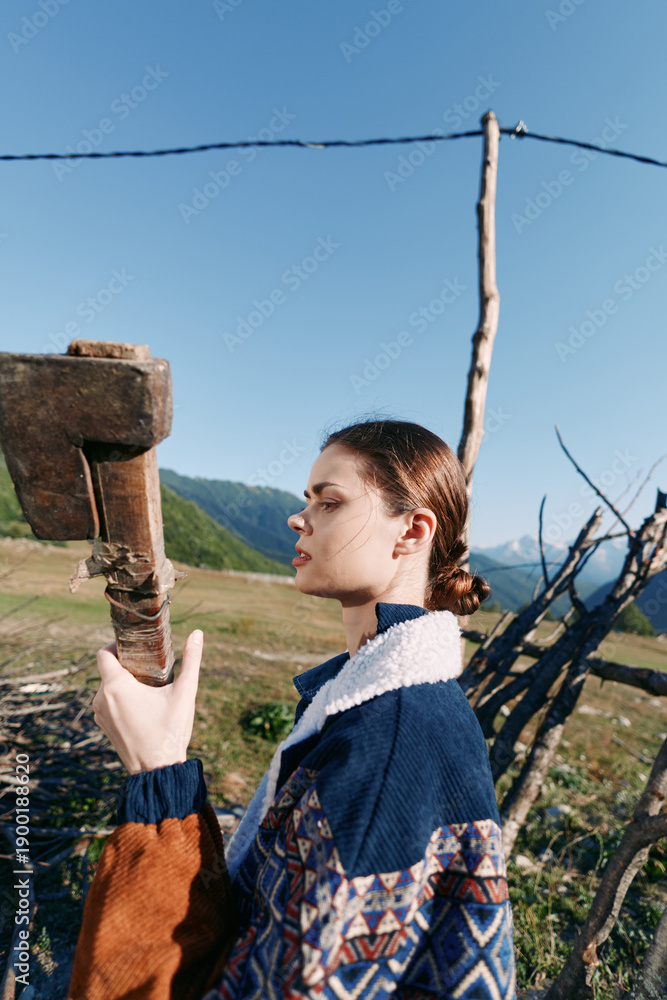 Fototapeta premium Woman portrait in countryside by a wooden fence, wearing patterned sweater in profile against mountains. Rural outdoors scene with sky, grass and serene seasonal mood.