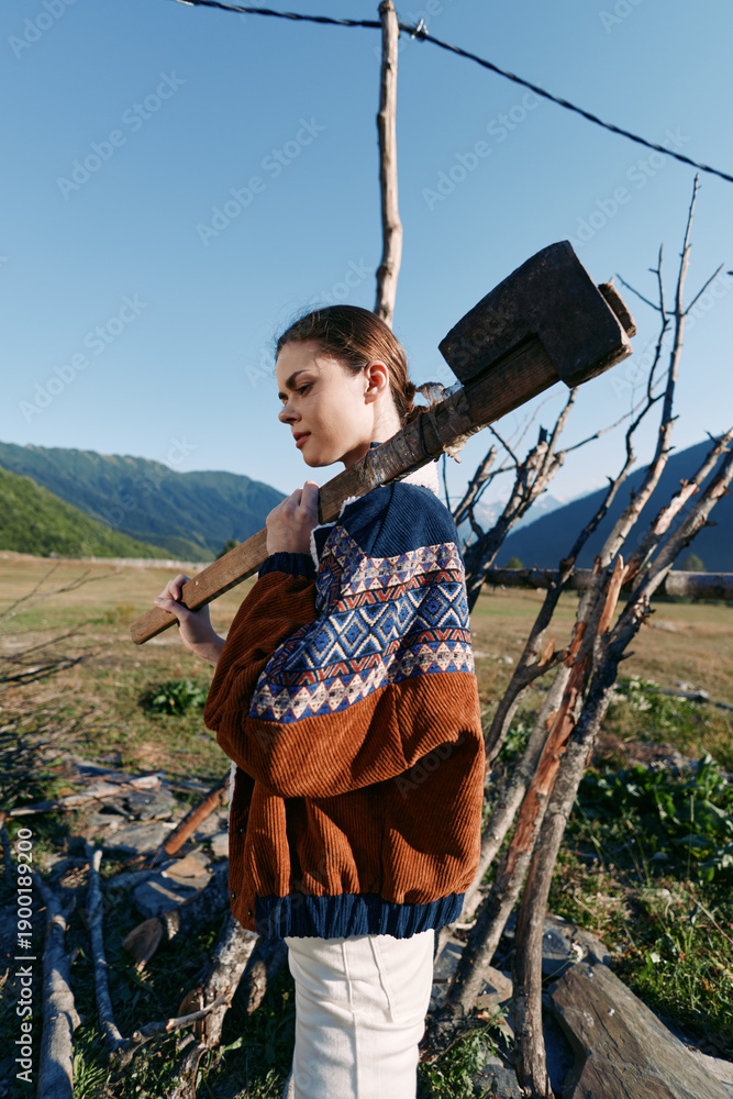 Fototapeta premium Woman axe wood countryside mountains sweater outdoor rural profile carrying large splitting axe over shoulder near wooden fence and stacked logs in mountain meadow landscape.