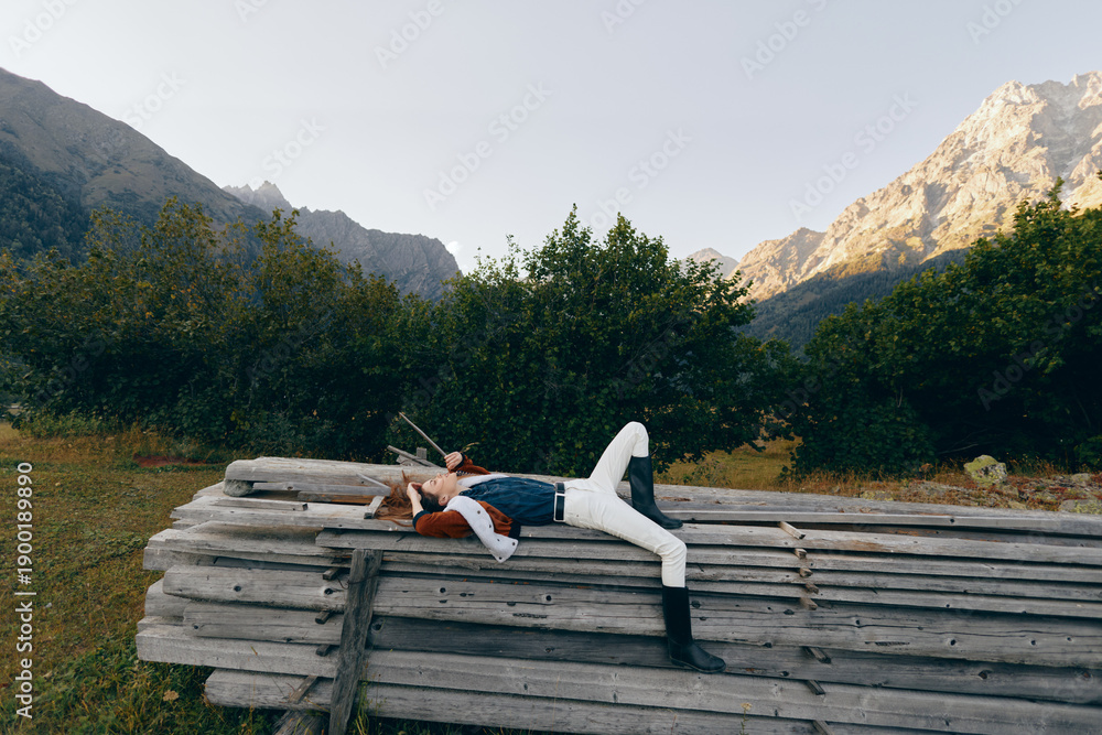 Fototapeta premium Woman resting on stacked wood beams in an orchard, surrounded by trees and mountains, outdoors relaxation scene with casual denim and boots, peaceful summer landscape.