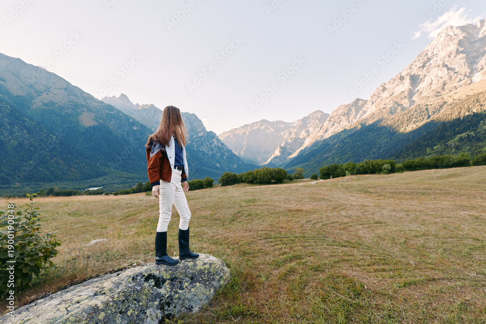Fototapeta premium Hiker woman mountains backpack meadow hiking landscape nature. Female traveler stands on a rock overlooking an alpine valley and rugged peaks, wearing boots and jacket for outdoor adventure.