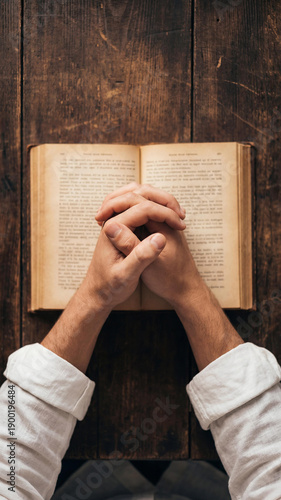 A top-down view of a person's hands clasped in prayer or contemplation over an open, aged book on a rustic wooden table. The scene evokes a sense of deep thought, study, or spiritual reflection