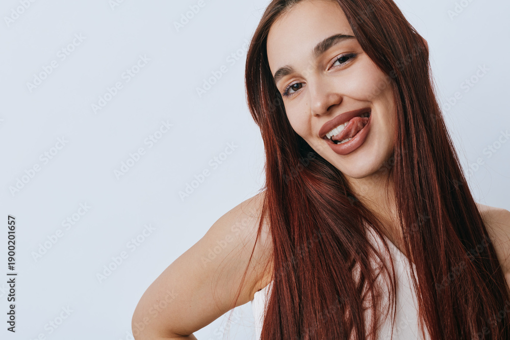 Obraz premium Young woman with long brown hair smiling playfully and sticking out her tongue against a plain light background. Casual portrait with natural expression and relaxed pose.