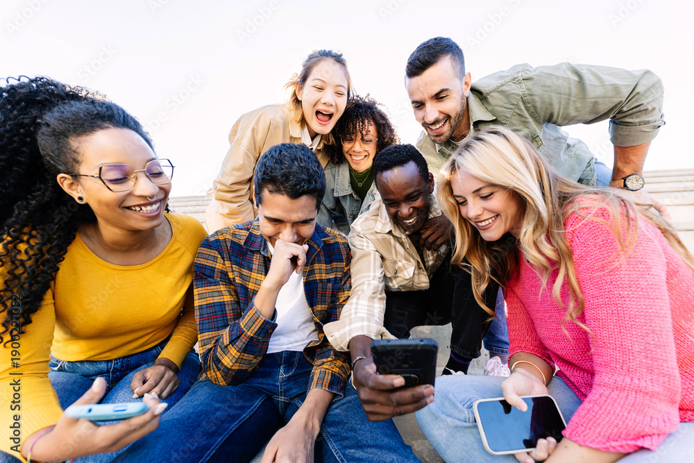 Fototapeta premium Multiracial group of young students enjoying funny content on a smartphone while relaxing together outdoors. Youth community and friendship lifestyle.