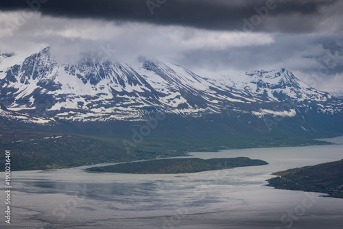 Dark storm clouds hang low over steep snow covered mountains dropping into a calm fjord near Tromsø in Northern Norway. Patches of spring greenery on the lower slopes contrast with the rugged peaks