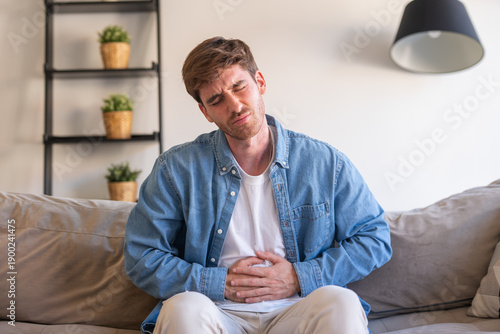 Tableau sur toile A sick young man sits at home holding his stomach in pain, suffering from abdomi