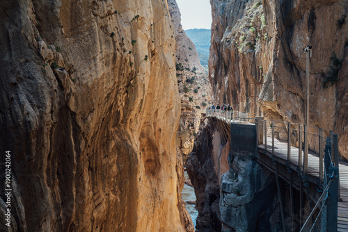 Group of tourists walking on narrow boardwalk along steep limestone cliffs in deep river gorge during Caminito del Rey hiking tour in Malaga Spain while enjoying scenic mountain canyon landscape.