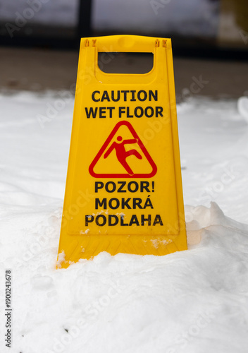 Yellow caution sign standing on a wet floor partially covered with snow, warning of slippery surface. Concept of winter safety, hazard prevention, risk awareness and cold weather conditions indoors.