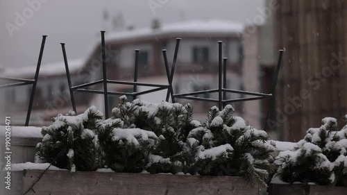 Snow blankets evergreen branches in wooden planters during a light snowfall in Cortina. Metal barstool frames stand inverted against a blurred backdrop of alpine buildings under a grey winter sky.