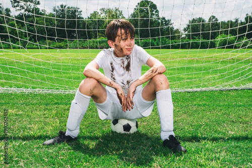 Youth soccer player sitting on a ball in the goal covered with mud