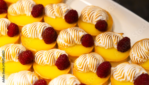 Bite size lemon meringue tarts with fresh raspberry accent  on each in close up of multiple sweet pastries placed on rectangular white plate