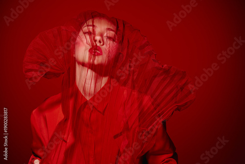 Obraz na plátně Artistic beauty portrait of a fashion model with red veiled fabric covering her face in a creative, dramatic red background studio setting