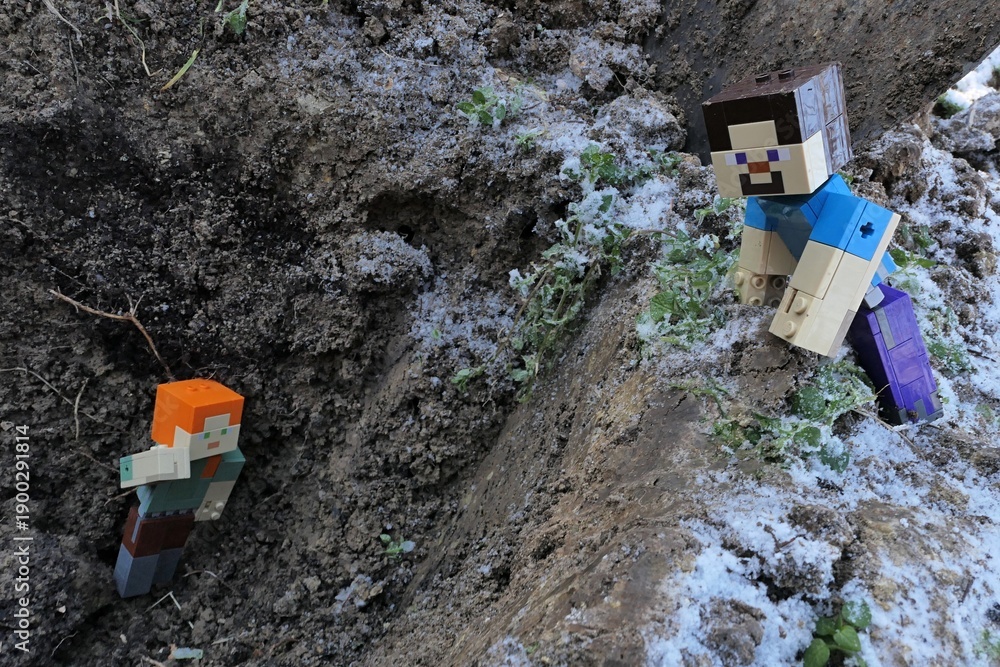 Fototapeta premium Minecraft action figure of smiling Steve spotted main female character figure of Alex on bottom of deep dug out garden pit. Winter half frozen garden soil around. 