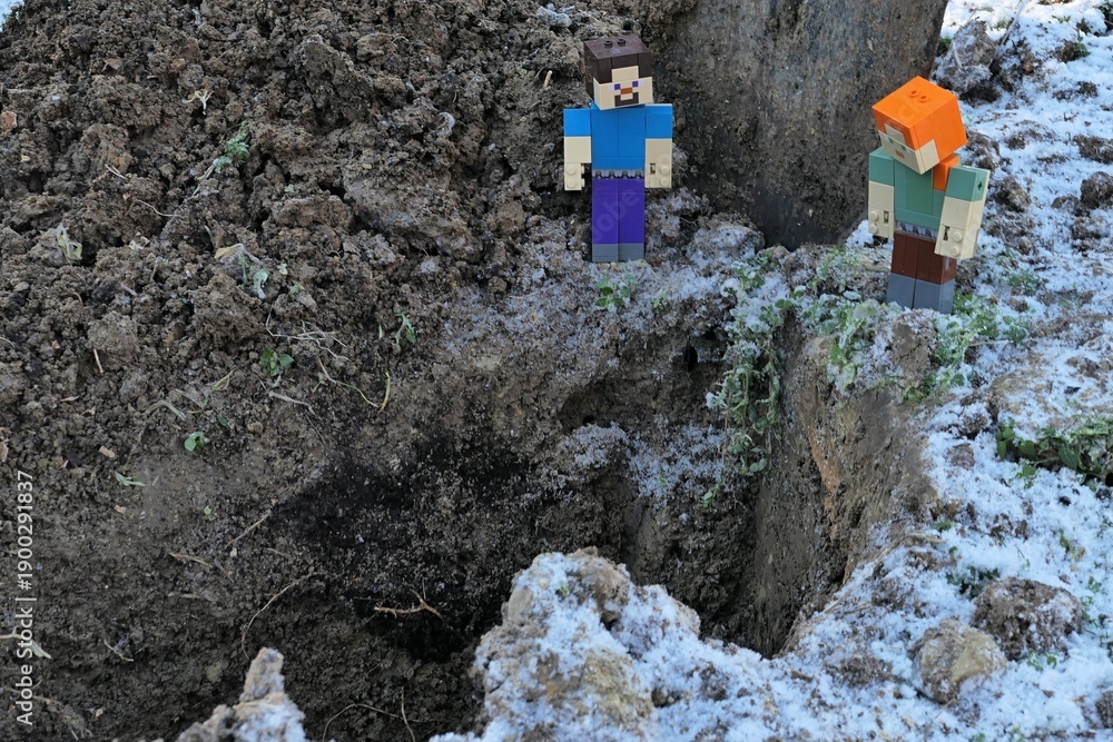 Obraz premium Minecraft action figures of Steve and Alex standing on the corner of deep garden pit, frozen soil and some snow visible around. 