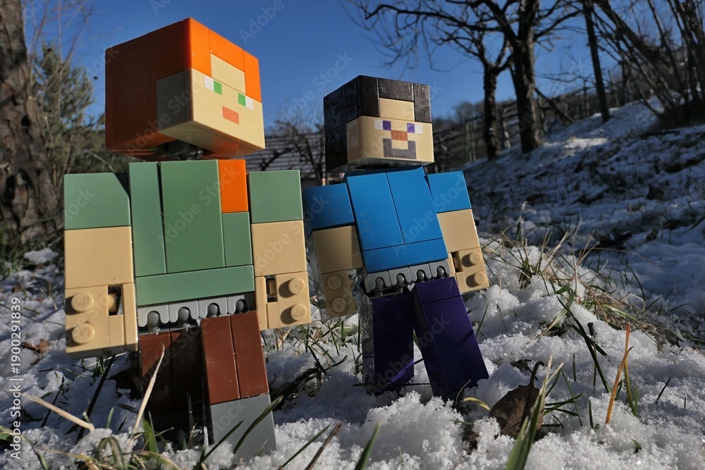 Fototapeta premium Minecraft action figures of smiling Steve and Alex on a walk in winter garden, first snow visible on the lawn. Naked fruit trees and shrubs as well as clear blue skies in background. Afternoon sun. 
