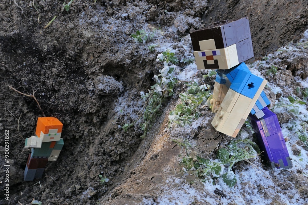 Fototapeta premium Minecraft action figure of Steve watching main female characters Alex fallen into dug garden hole, traces of winter snow and frozen soil around. 
