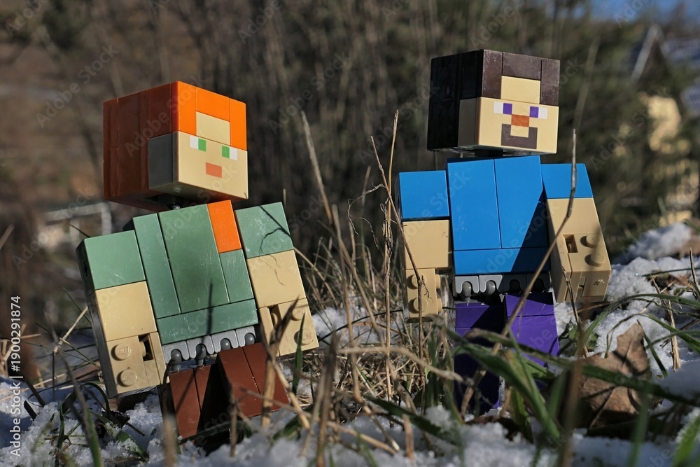 Fototapeta premium Minecraft figures of Steve and Alex walking among stems of grass of garden lawn, partially covered with first winter snow, sunlit by afternoon sunshine. 