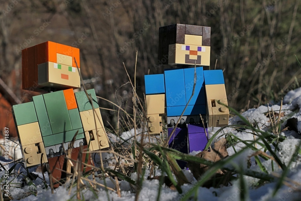 Fototapeta premium Minecraft figures of Steve and Alex on a walk among stems of grass of garden lawn, partially covered with first winter snow, sunlit by afternoon sunshine. 