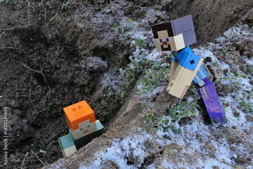 Fototapeta premium Minecraft figure of main character Steve looking at her female friend Alex trying to get out from a pit in frozen winter garden, slightly covered with first snow. 