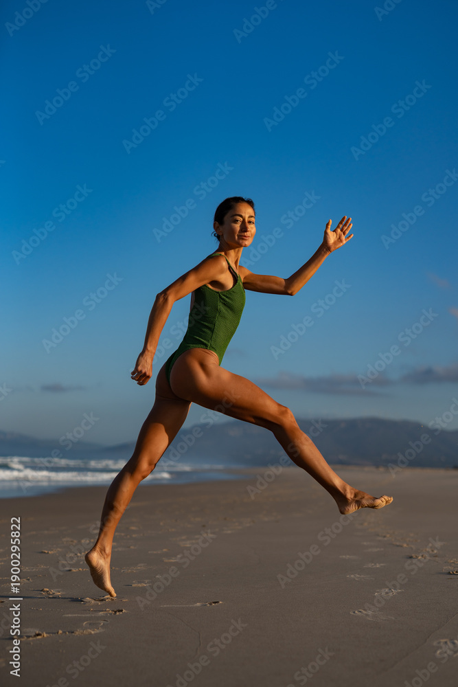 Fototapeta premium Fit young woman in green swimsuit mid-air as she jumps gracefully along the beach at sunrise, showing strength, energy, and freedom under a clear blue sky.
