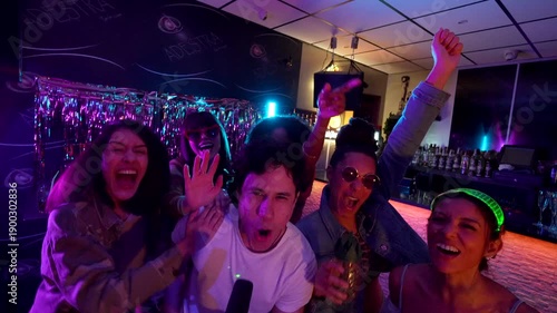 Happy young man singing into a microphone during a karaoke night, with his cheerful, diverse group of friends dancing, laughing, and singing along in a vibrant, neon lit bar setting