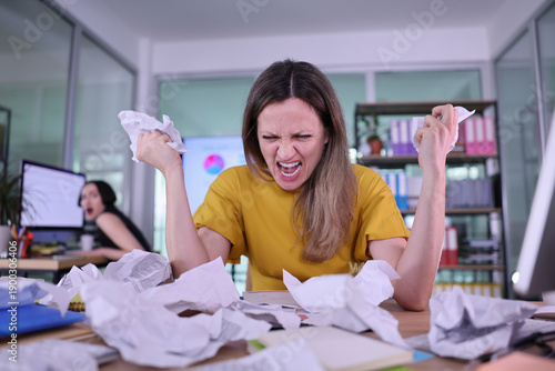 Fotografie A young woman in a yellow shirt expresses her frustration among a clutter of crumpled papers