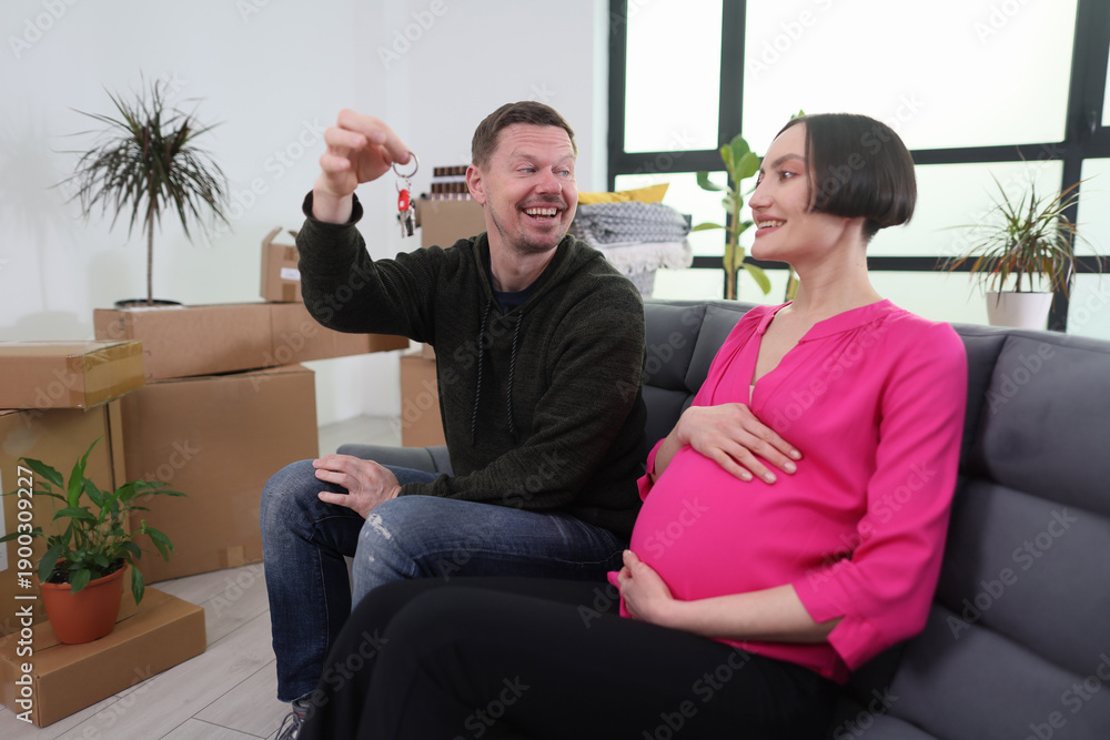 Obraz premium In a bright living room, a happy couple shares a joyful moment as they sit on a stylish couch. The man holds up house keys while the pregnant woman smiles, cradling her baby bump.