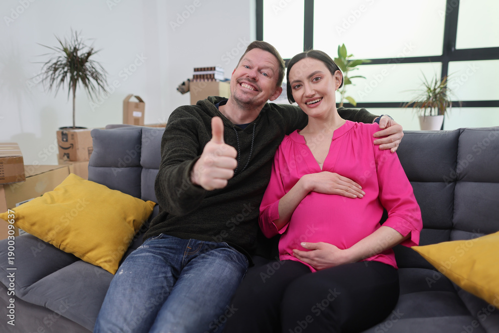 Obraz premium A happy couple sits on a comfortable couch, sharing smiles and laughter as they embrace the journey of pregnancy. Boxes and plants surround them, creating a cozy environment.