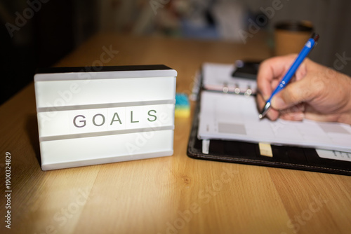 Person writing goals in planner with lightbox on desk