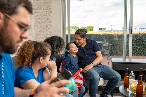 Girl wearing a USA flag looks at the camera while family and friends enjoy watching football soccer at home. Sports togetherness, cultural pride, joyful moments, friendship, casual home viewing