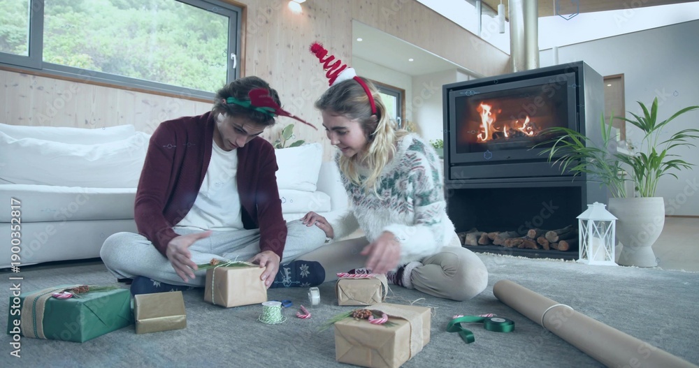 Obraz premium Wrapping women wearing festive headbands and sweaters at home, handling kraft gifts and ribbons