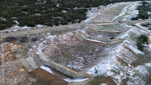 Aerial shot looking down at tiered concrete drainage basins or erosion control steps on a snowy hillside surrounded by low green bushes in winter